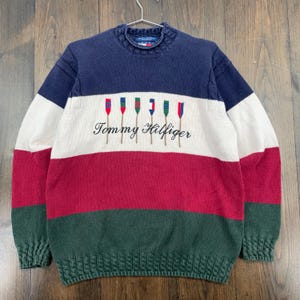 May include: A navy blue, white, red, and green Tommy Hilfiger sweater. The sweater features a color block design with the brand name and a rowboat graphic embroidered on the front. The sweater has a ribbed collar, cuffs, and hem.