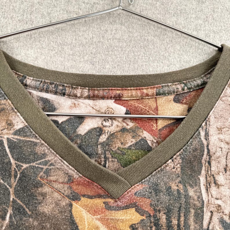 Pode incluir: Uma t-shirt com decote em V apresentando um padr&atilde;o de camuflagem realista de solo florestal com folhas ca&iacute;das e texturas de casca de &aacute;rvore em tons de castanho, bege e verde. A pe&ccedil;a tem um acabamento canelado verde-azeitona no decote e um cabide.