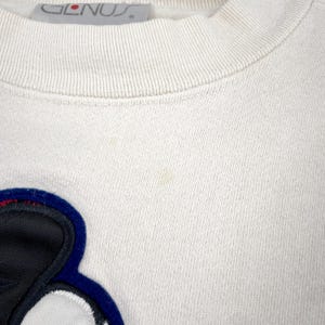 May include: Cream-colored sweatshirt with a crew neck and a large, colorful applique. The applique features a black, white, and blue design. The brand name "GLENUS" is visible on the tag.