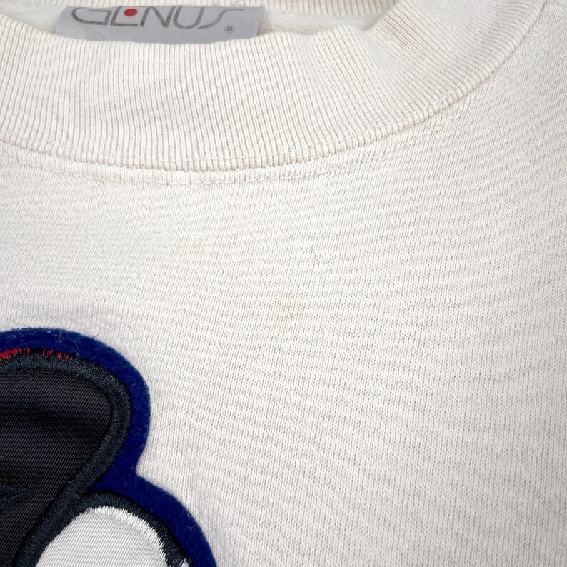 May include: Cream-colored sweatshirt with a crew neck and a large, colorful applique. The applique features a black, white, and blue design. The brand name "GLENUS" is visible on the tag.