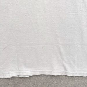 May include: Close-up of a white t-shirt. The fabric is slightly wrinkled, with a visible seam at the bottom edge. A small, faint pink stain is present on the shirt's surface. The shirt appears to be made of a soft, lightweight material.