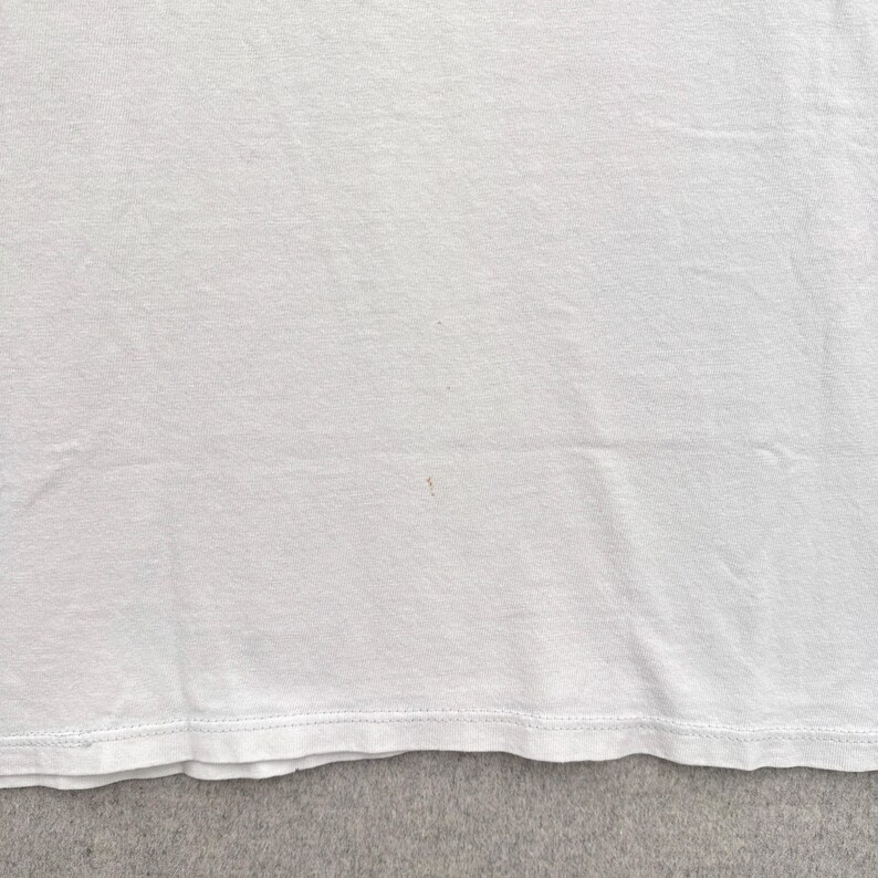 May include: Close-up of a white t-shirt. The fabric is slightly wrinkled, with a visible seam at the bottom edge. A small, faint pink stain is present on the shirt's surface. The shirt appears to be made of a soft, lightweight material.