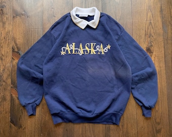 May include: A navy blue sweatshirt with a white collar and the word "ALASKA" embroidered in gold with stars on either side.