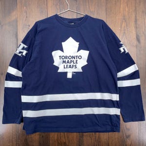 May include: Navy blue long-sleeve shirt featuring the Toronto Maple Leafs logo in white. The shirt has white stripes across the chest and sleeves, and the team logo is also on the sleeves. The shirt is hanging on a metal hanger.