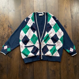 May include: A white, green, and navy blue argyle cardigan with a v-neck and button closure. The cardigan has two pockets on the front.