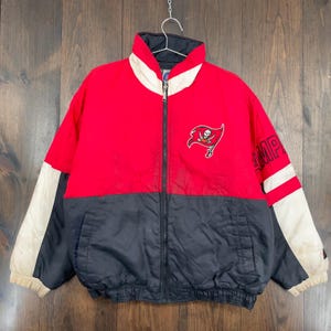 May include: A red, black, and white zip-up jacket featuring the Tampa Bay Buccaneers logo on the chest. The jacket has a high collar and the word "CHAMP" is printed on the sleeve. The jacket is suspended from a metal hook.