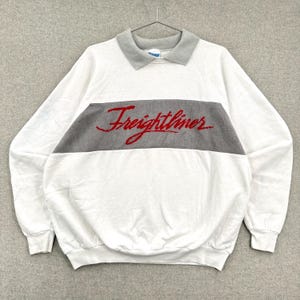 Op de afbeelding: Wit sweatshirt met een grijze kraag en een grijze horizontale streep over de borst. Het woord "Freightliner" is in rood script geborduurd over de grijze streep. De sweatshirt heeft lange mouwen en een ronde hals.