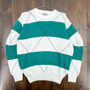 Op de afbeelding: Witte en teal argyle trui met horizontale teal strepen. De trui heeft een ronde hals en lange mouwen. Het argyle patroon is gemaakt met kleine zwarte steken. De trui hangt aan een metalen hanger.