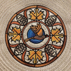 May include: Embroidered round design featuring a stylized bird flying through a circle of leaves and pine cones. The design is in shades of brown, orange, yellow, and blue.