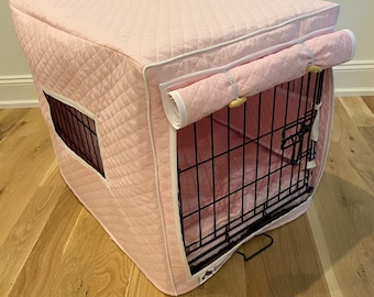 May include: A pink quilted dog crate cover with a white trim and a roll-down flap. The cover has a small window for visibility.