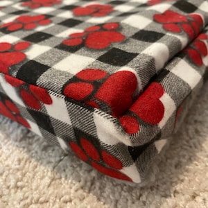 May include: Close-up of a red and black checkered dog bed with paw prints. The bed is made of soft, plush fabric and has a black border.