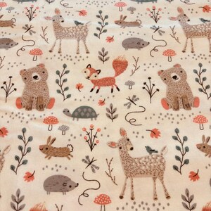 May include: A cream-coloured fabric with a repeating pattern of woodland creatures. The design includes deer, foxes, bears, rabbits, hedgehogs, and turtles, along with mushrooms, leaves, and sprigs. The illustrations are in shades of brown, orange, and grey.
