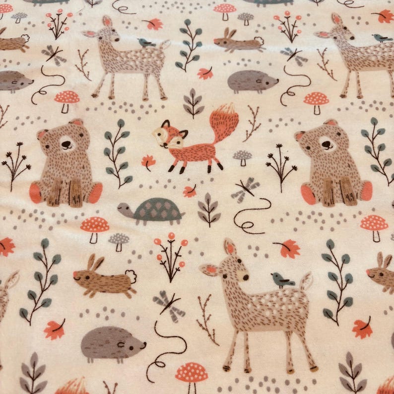 May include: A cream-coloured fabric with a repeating pattern of woodland creatures. The design includes deer, foxes, bears, rabbits, hedgehogs, and turtles, along with mushrooms, leaves, and sprigs. The illustrations are in shades of brown, orange, and grey.
