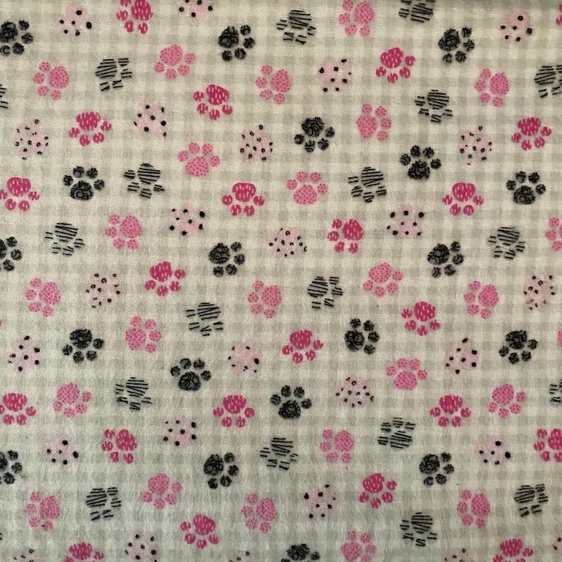 May include: A fabric featuring a repeating pattern of pink and black paw prints and small dots on a light grey checkered background. The paw prints vary in size and direction, creating a playful design.