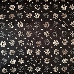 May include: Black fabric featuring a repeating pattern of white and silver paw prints and small white and grey hearts. The paw prints vary in style, some with solid fills and others with striped details.