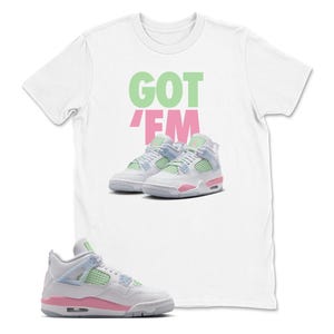 May include: White t-shirt with the text "GOT EM" in green and pink. The shirt features an image of white, pink, and light blue trainers. A single trainer is shown in the foreground.