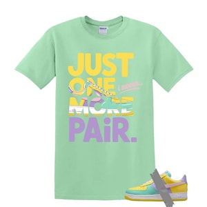 May include: A light green t-shirt with the text "JUST ONE MORE PAIR" in yellow and purple. The graphic features a sneaker design. A matching sneaker is taped to the bottom of the shirt.