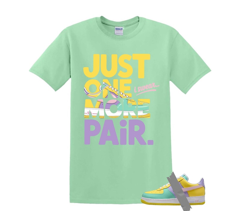 May include: A light green t-shirt with the text "JUST ONE MORE PAIR" in yellow and purple. The graphic features a sneaker design. A matching sneaker is taped to the bottom of the shirt.