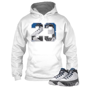 May include: White hoodie with a blue and white "23" graphic on the front. The hoodie is paired with a pair of white, black, and blue high-top sneakers. The sneakers have a lace-up design and a rubber sole.