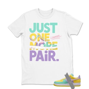 May include: White t-shirt with the text "JUST ONE MORE PAIR" in teal, yellow, purple, and pink. A colorful sneaker is shown at the bottom right. The sneaker is yellow, green, purple, and pink.