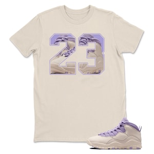 May include: A cream-colored t-shirt featuring the number 23 in a light purple and cream design. A pair of light purple and cream sneakers are in the lower right corner.