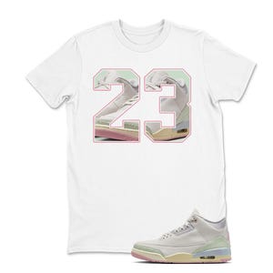 May include: White t-shirt with a large "23" graphic in pink, showcasing a sneaker design. A matching sneaker is positioned below the shirt. The sneaker is light gray with pastel accents of pink, green, and yellow.