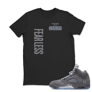 May include: Black t-shirt with the word "FEARLESS" printed vertically in gray on the left side. The shirt also features the word "fearless" and a quote in gray. A gray and blue sneaker is in the foreground.