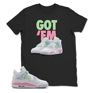 May include: Black t-shirt with the words "GOT 'EM" in green and pink. The shirt features a graphic of white, pink, and light blue trainers. A pair of matching trainers is in the foreground.