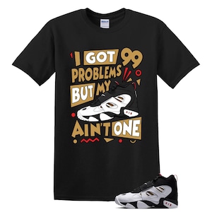 May include: A black short-sleeve t-shirt featuring a graphic of a white, black, and red sneaker. The text reads I GOT 99 PROBLEMS BUT MY AIR AIN'T ONE, arranged in gold and white blocks with red and black geometric accents surrounding the sneaker.