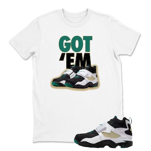 May include: White t-shirt with the text "GOT 'EM" in green and black. The shirt features a graphic of black, white, and green athletic shoes. A single shoe is shown at the bottom right.