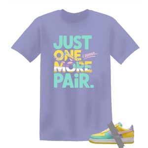 May include: A light purple t-shirt with the text "JUST ONE MORE PAIR. i swear..." in teal, yellow, and white. The graphic features a colorful sneaker design. A matching sneaker is visible at the bottom.
