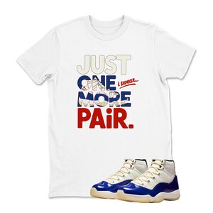 May include: White t-shirt with the text "JUST ONE MORE PAIR" in red, white, and blue. Below the shirt are a pair of white and blue sneakers.
