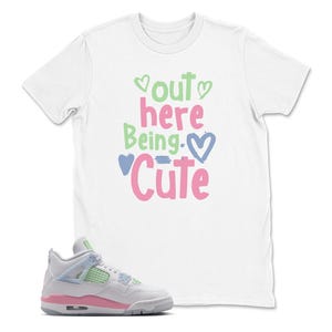 May include: White t-shirt with the phrase "Out Here Being Cute" in green, pink, and blue. A pair of white, pink, and light blue sneakers are in the foreground.