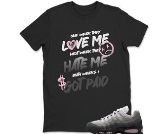 May include: A black t-shirt featuring white and pink text that reads: ONE WEEK THEY LOVE ME NEXT WEEK THEY HATE ME BOTH WEEKS I GOT PAID, with a pink heart, a pink sad face, and a pink dollar sign. A grey and black sneaker is positioned below.