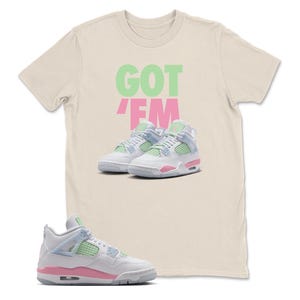 May include: A cream-coloured t-shirt with the words "GOT EM" in green and pink. The shirt features a graphic of white and pink trainers with light blue and green accents. The trainers are a popular athletic shoe style.