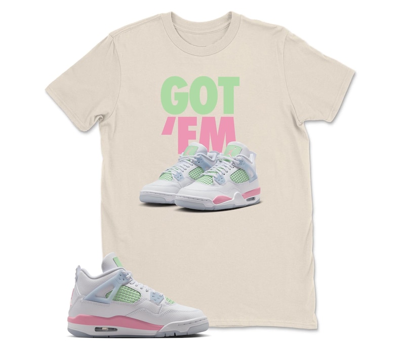 May include: A cream-coloured t-shirt with the words "GOT EM" in green and pink. The shirt features a graphic of white and pink trainers with light blue and green accents. The trainers are a popular athletic shoe style.