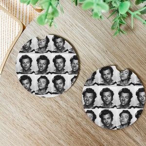 May include: Two round, white coasters featuring a black and white photograph of a man's face repeated in a grid pattern. The coasters are on a wooden surface.