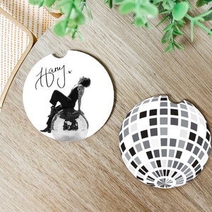 May include: Two round white coasters. One features a black and white photo of a person sitting on a disco ball with the signature "Harry." The other coaster has a black and white design of a disco ball.