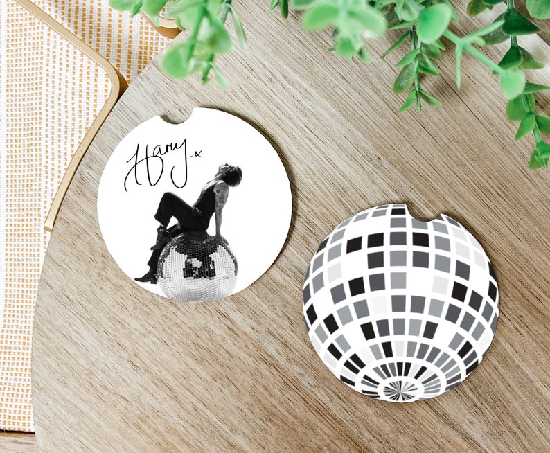 May include: Two round white coasters. One features a black and white photo of a person sitting on a disco ball with the signature "Harry." The other coaster has a black and white design of a disco ball.