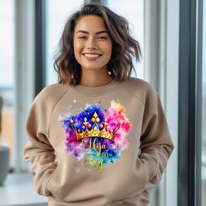 May include: A tan sweatshirt with a watercolor design of a gold crown and the text "Hija del Rey" in a purple and gold font.