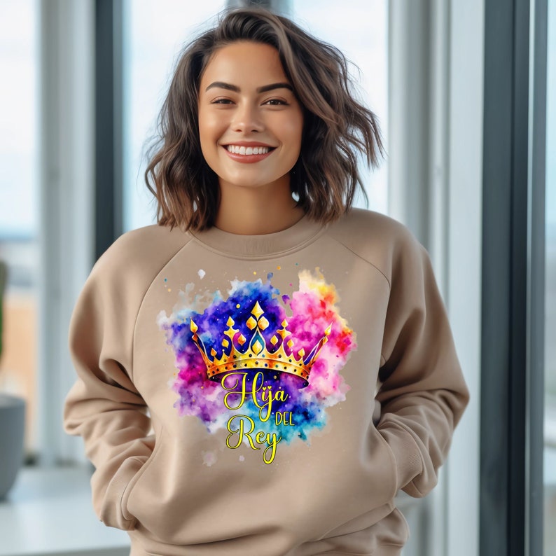 May include: A tan sweatshirt with a watercolor design of a gold crown and the text "Hija del Rey" in a purple and gold font.
