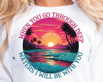Puede incluir: Sudadera blanca con un diseño gráfico circular que presenta una puesta de sol sobre una playa con palmeras. El texto "WHEN YOU GO THROUGH DEEP WATERS I WILL BE WITH YOU" rodea la imagen. El texto "ISAIAH 43:2" también está incluido en el gráfico.