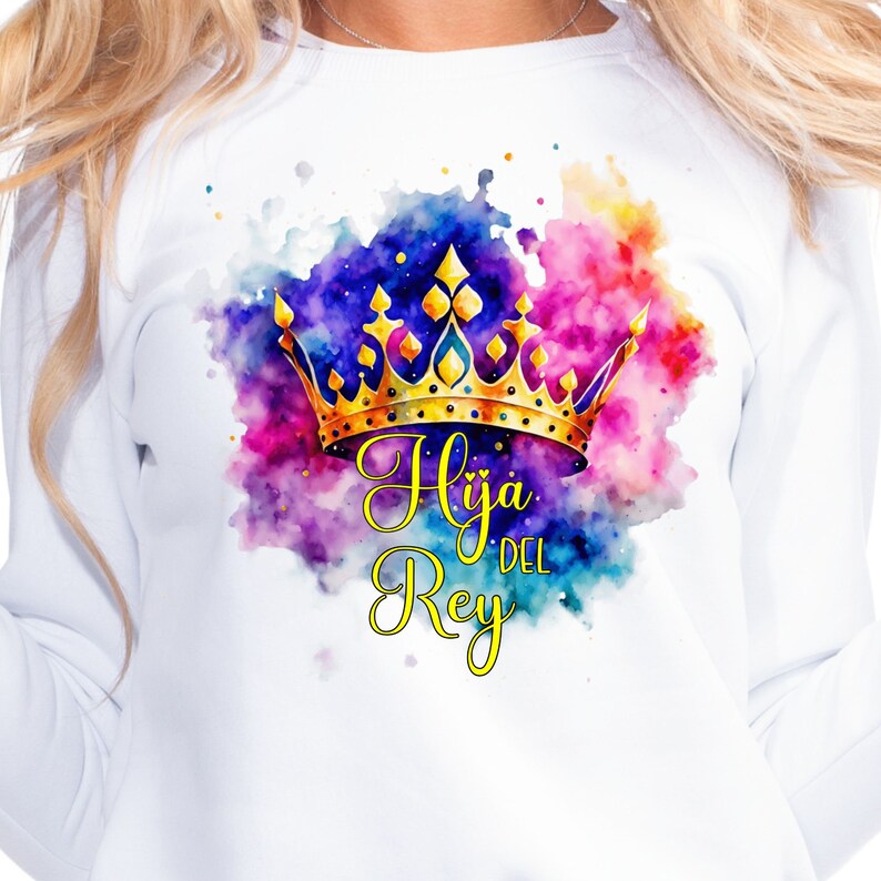 May include: White sweatshirt with a watercolor design of a gold crown and the text "Hija del Rey" in yellow.
