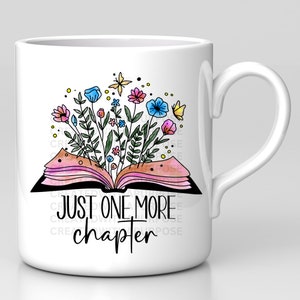 May include: White ceramic mug featuring a colorful illustration of an open book with flowers and butterflies. The text "JUST ONE MORE chapter" is printed below the book. The mug has a curved handle.