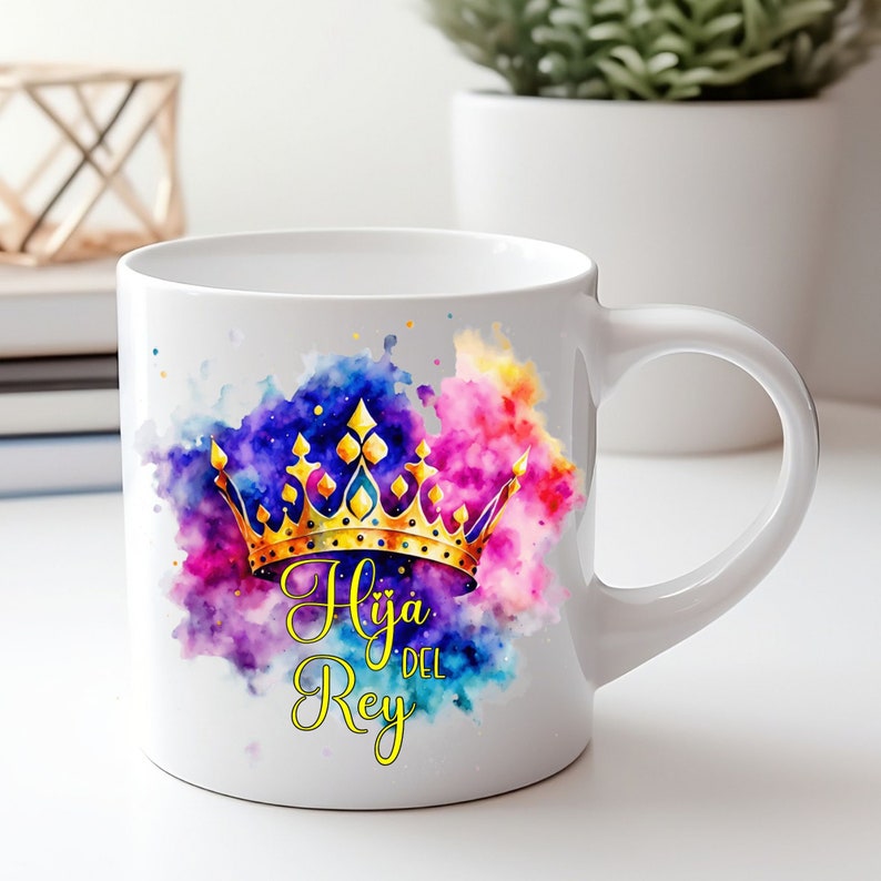 May include: White ceramic mug with a colorful watercolor design featuring a gold crown and the text "Hija del Rey" in yellow.