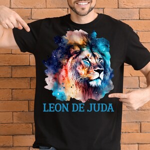 May include: A black t-shirt with a colorful watercolor illustration of a lion's head. The lion's mane is a mix of blue, orange, and yellow. The background is a galaxy-like pattern with stars. The text "LEON DE JUDA" is printed in blue below the lion's head.