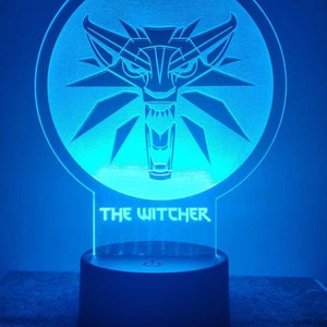 May include: A blue LED night light with a white wolf head design and the text "THE WITCHER" on the front. The light is on a black base with a power switch.