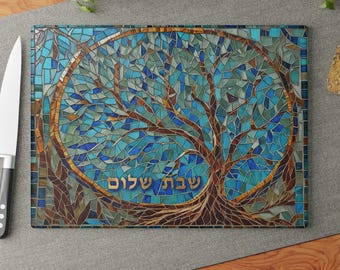 May include: A rectangular glass cutting board featuring a mosaic-style tree design in shades of blue, teal, and brown. The tree is centered within a circular frame, and the Hebrew text "שבת שלום" is printed in gold lettering at the base.