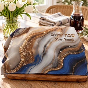 May include: A rectangular fabric cover featuring a marbled pattern in shades of blue, white, and brown with gold glitter accents. The text "Shabbat Shalom" in Hebrew and "Stein Family" in English is printed on the right side of the fabric.