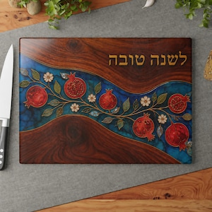 May include: A rectangular cutting board featuring a dark wood grain finish with a central wavy blue marbled band. The design includes a vine with red pomegranates, green leaves, and white flowers. The Hebrew text "לשנה טובה" is printed in gold.